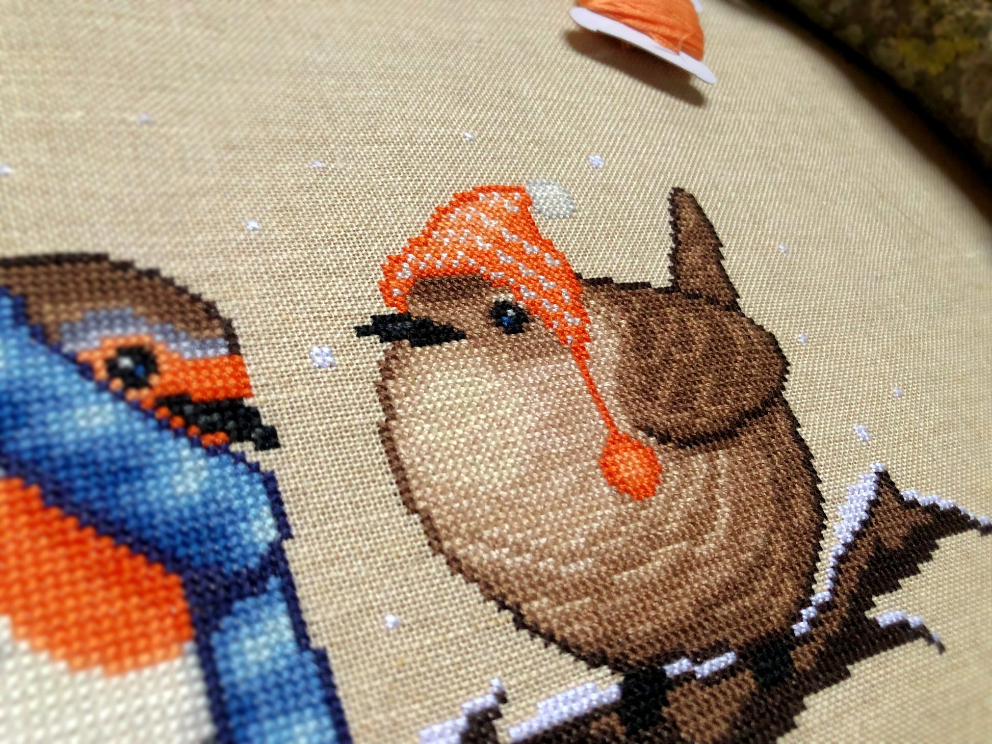 Closeup of Winter Birds cross stitch pattern. Image is zoomed in on the wren. He is very cute and round. The wren is wearing an orange, striped hat with pompoms, and looks very warm. The colors of his feathers are brown and white.
