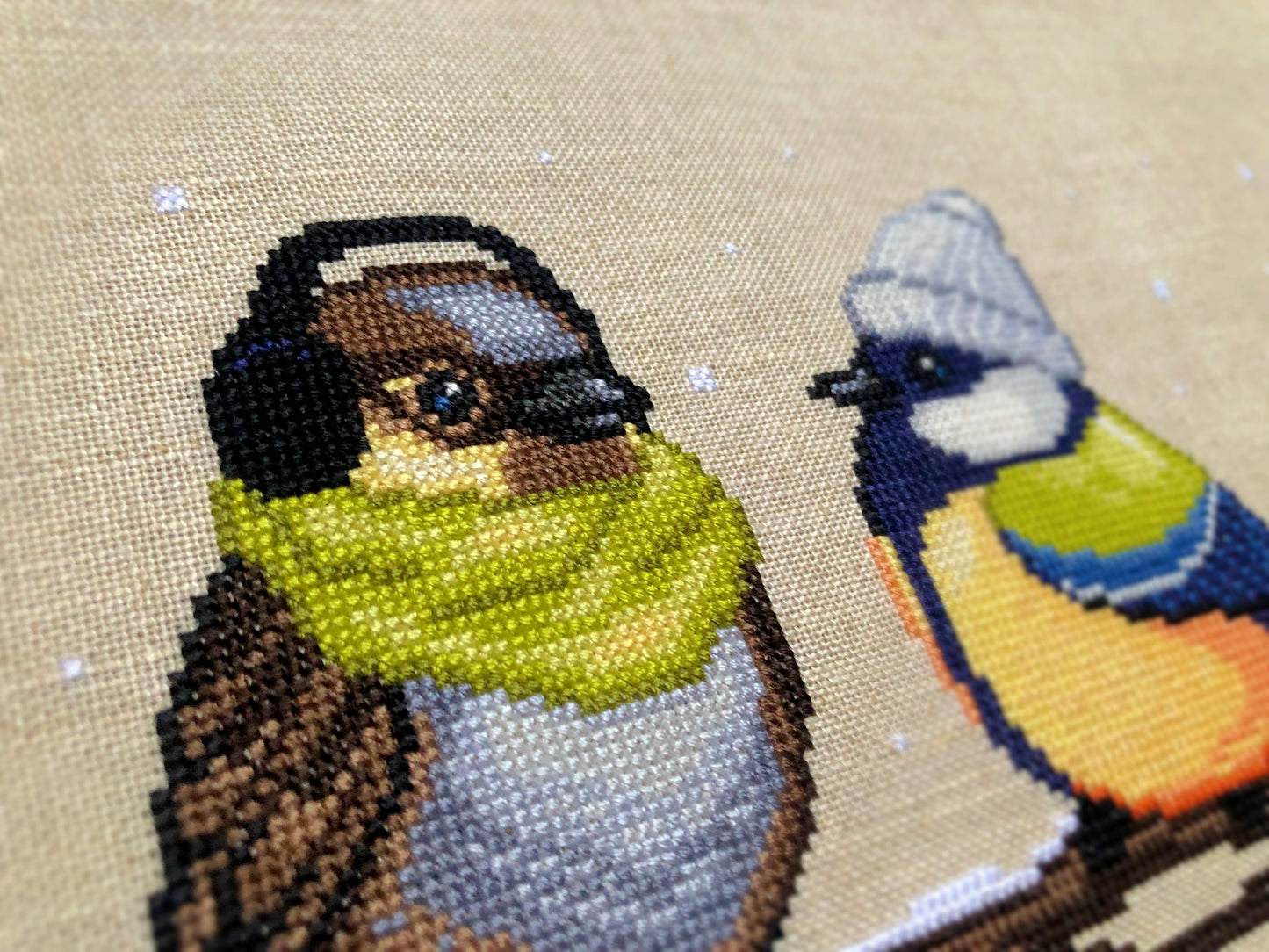 Closeup of Winter Birds cross stitch pattern. Image is zoomed in on the sparrow. He is a very cute bird that also looks very hip. The bird is wearing a green scarf and earmuffs, and looks very warm. The colors of his feathers are brown, beige, grey.